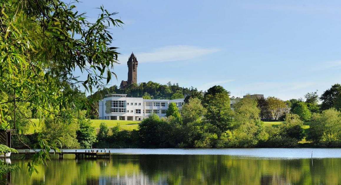 绿色的森林和平静的湖泊的照片, 后面有一座白色的建筑和一座高塔:斯特林的斯特林大学, 苏格兰.
